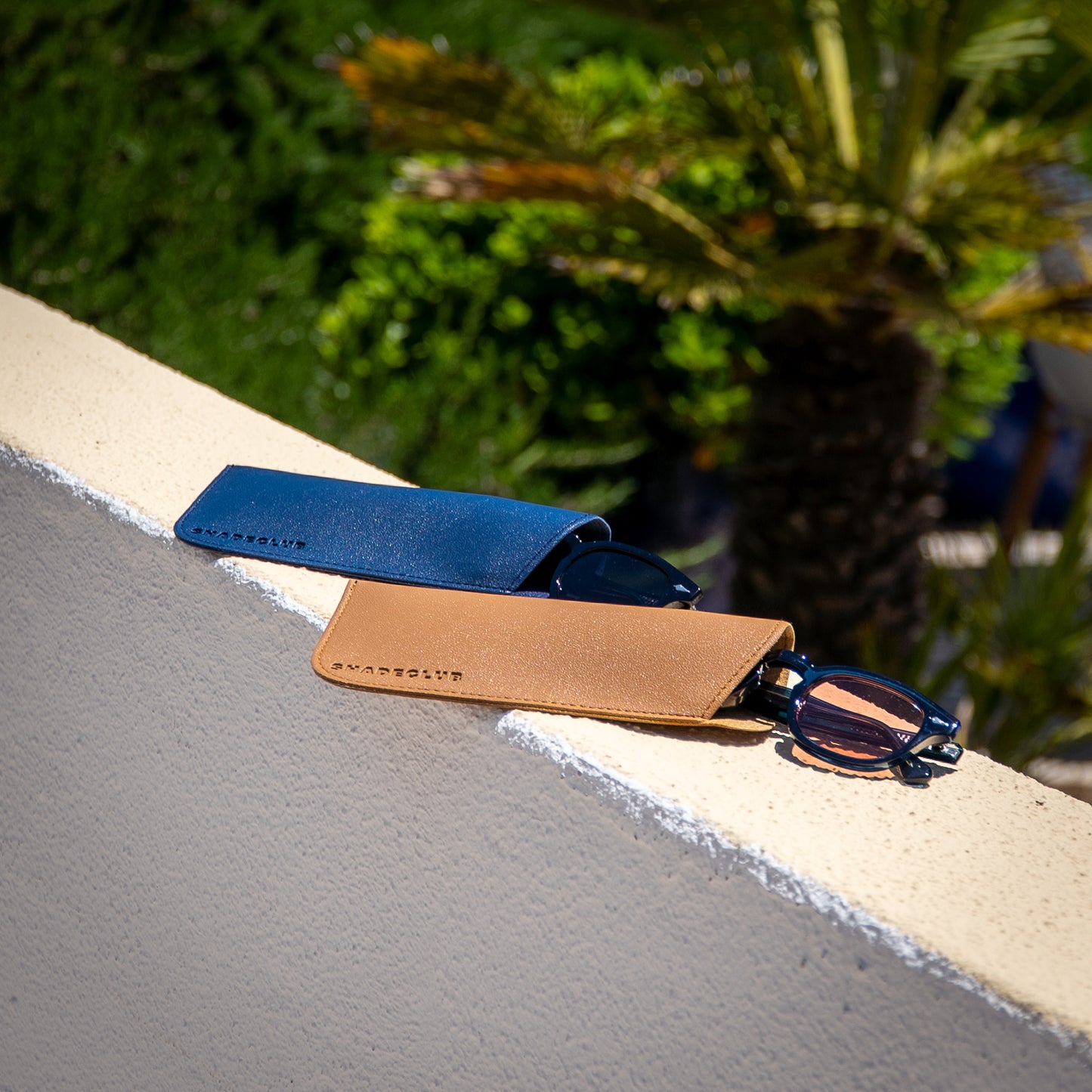 Monarch zonnebrillen half uit beige en blauwe hoes op stenen muurtje | luxe details in zomerse setting