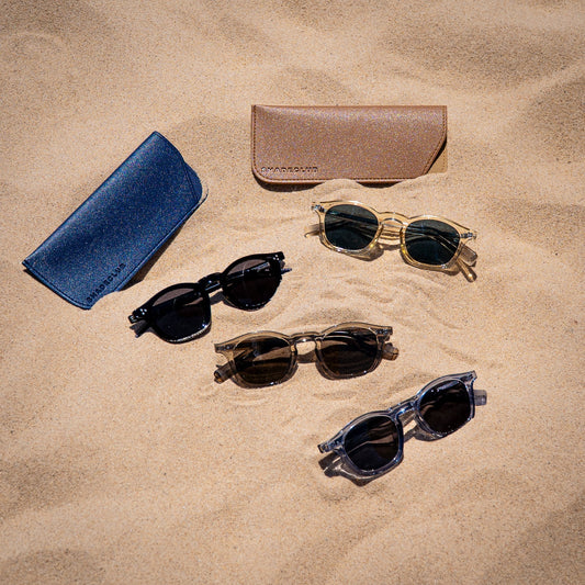 Shadeclub Collin zonnebrillen in alle kleuren naast elkaar in het zand met luxe hoesjes – stijlvolle en betaalbare zonnebrillen zomer 2025