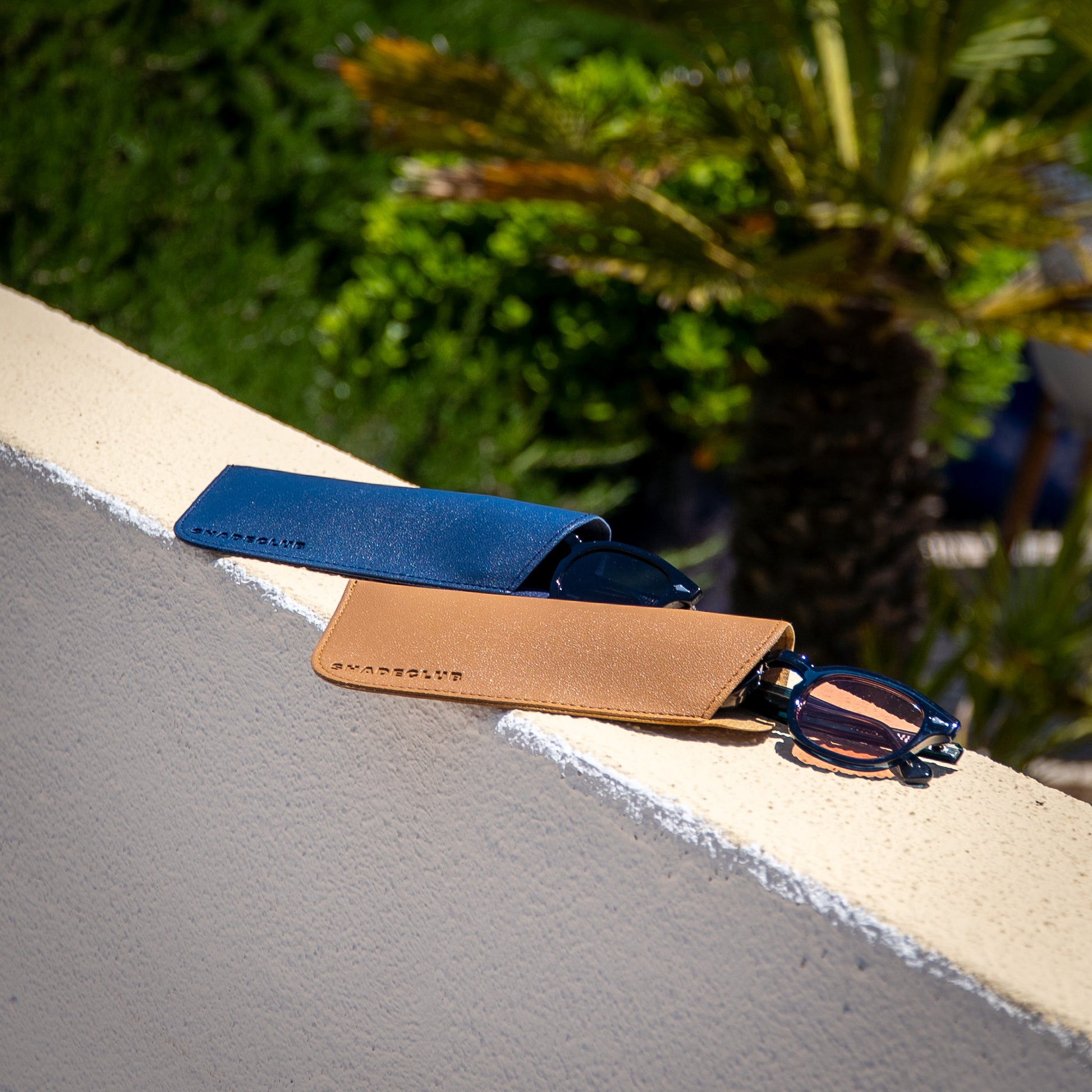 Monarch zonnebrillen half uit beige en blauwe hoes op stenen muurtje | luxe details in zomerse setting