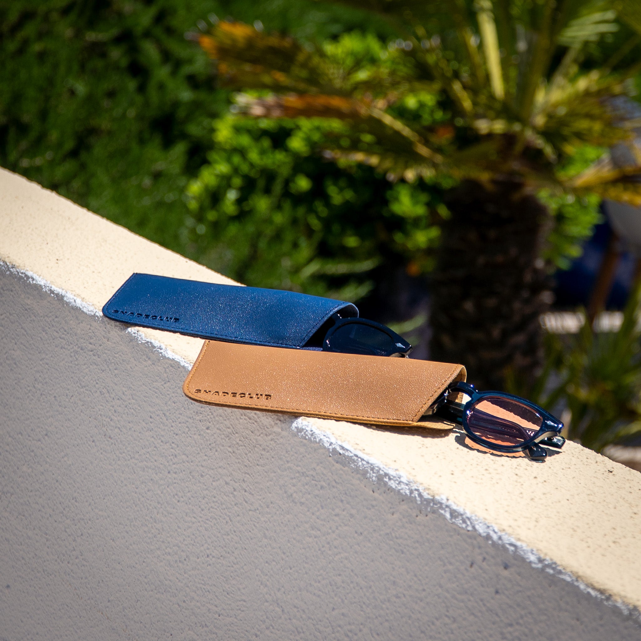 Monarch zonnebrillen half uit beige en blauwe hoes op stenen muurtje | luxe details in zomerse setting